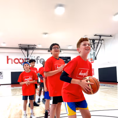 Group of boys wearing red hoopslife shirts standing in line on a basketball court, one holding a basketball and smiling.