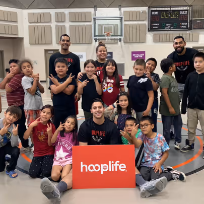 Group of children and three adults posing together in a gymnasium, with one adult holding an orange sign that says hooplife.