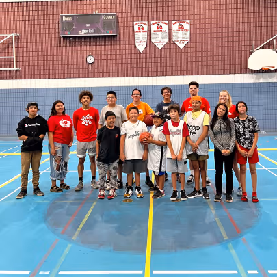 Group of children and teenagers standing together on an indoor basketball court, some wearing sports attire and holding a basketball.