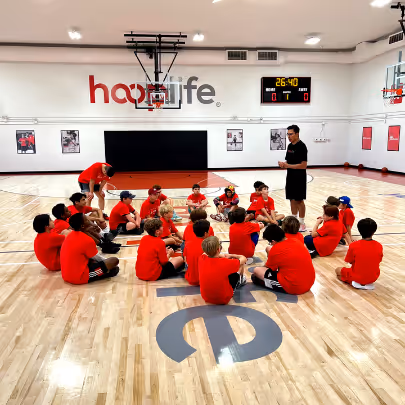 Basketball coach instructing a group of children in red shirts seated in a circle on an indoor basketball court.
