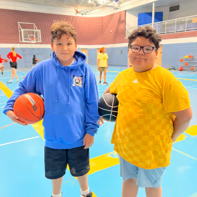 Two boys standing on an indoor basketball court, each holding a basketball and smiling at the camera.