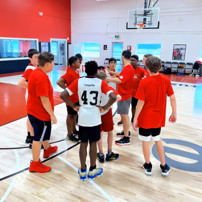 Group of boys, some wearing red shirts, standing in a basketball court during a team huddle or discussion.