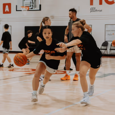 Two girls playing basketball indoors, one dribbling the ball while the other defends.