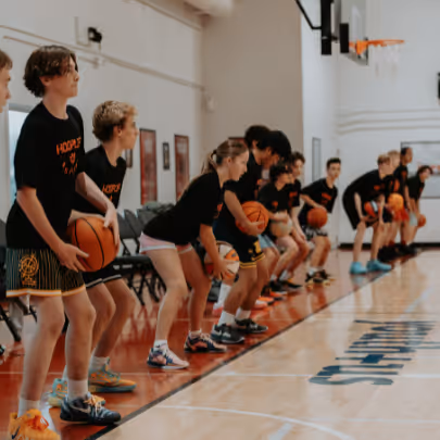A line of children in a gymnasium practicing basketball dribbling with balls in a bent-knee stance.