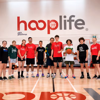 Group of nine young basketball players standing in a gym under a large HoopLife sign, some wearing red HoopLife shirts and one holding a basketball.