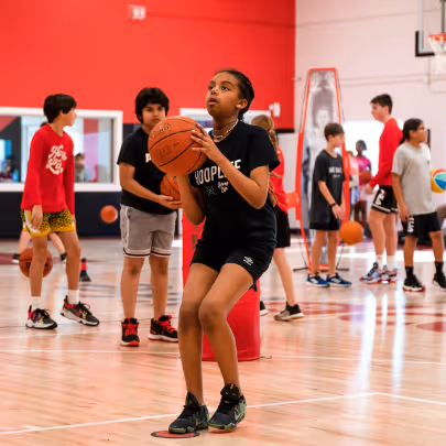 A young girl in black sportswear preparing to shoot a basketball in an indoor gym with other children playing basketball in the background.
