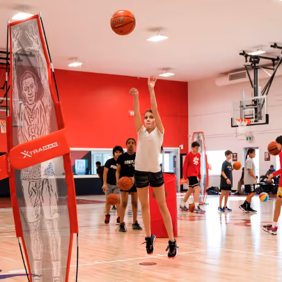 Young basketball player in mid-air shooting a basketball in an indoor gym with several other players in the background.