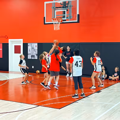 Youth basketball game with children reaching for the ball under the hoop on an indoor court with red walls.