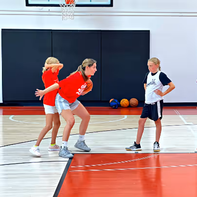 Three girls on an indoor basketball court practicing defense and offensive moves near the basket.