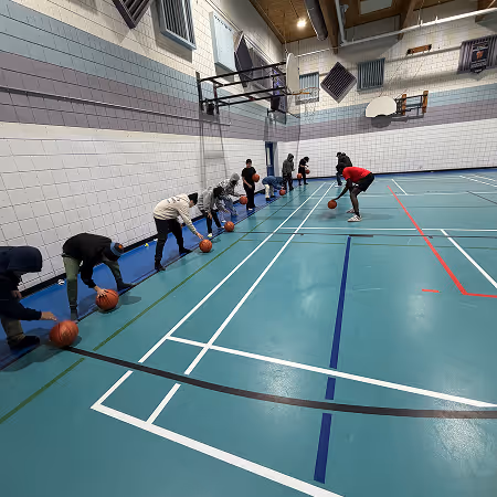 Children lined up in a gymnasium practicing basketball dribbling while a coach in a red shirt demonstrates.