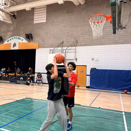 Two boys playing basketball indoors, one preparing to shoot a ball towards the hoop while the other defends.