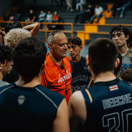 Basketball coach in an orange shirt speaking to his team during a timeout in an indoor gym.
