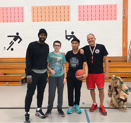 Two coaches and two boys standing on a gymnasium basketball court, one boy holding a basketball.