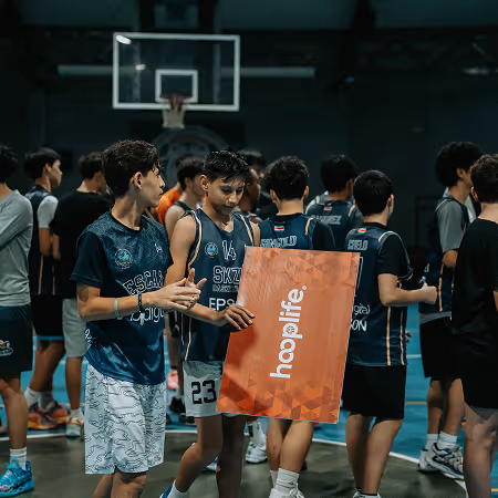 Young basketball players in uniforms standing on an indoor court, one holding a sign that reads 'hooplife.'