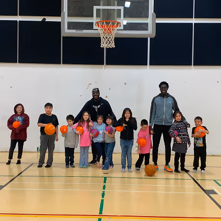 Two adult coaches standing behind a diverse group of children holding colorful basketballs in an indoor basketball court under a hoop.