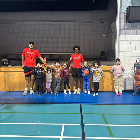 Two male coaches in red shirts standing with a group of young children in a gymnasium, some holding basketballs.